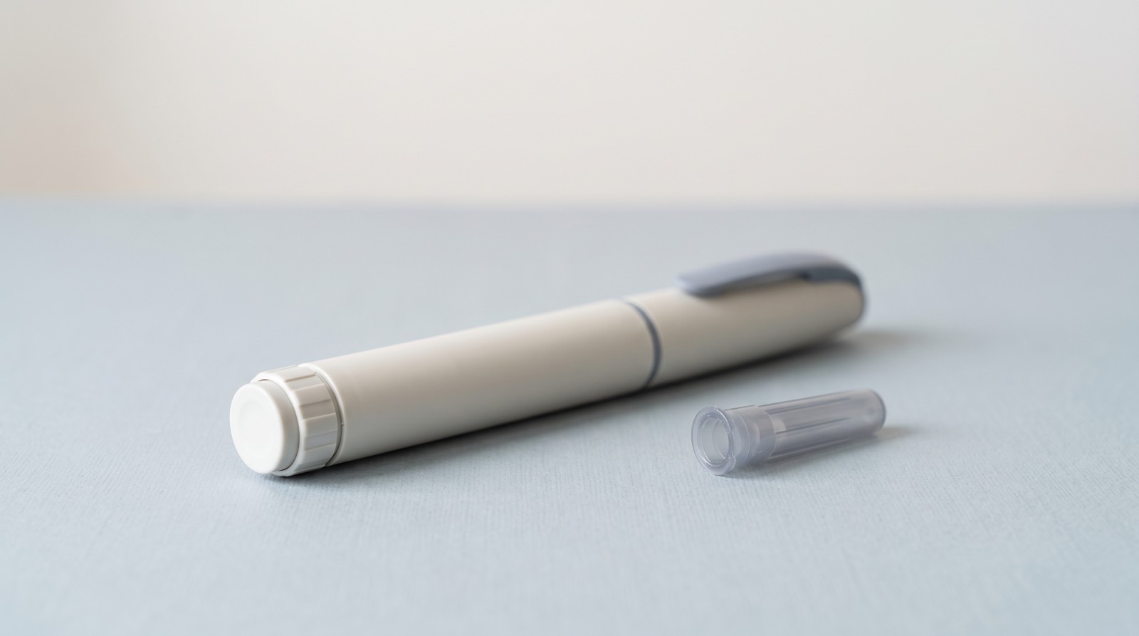 A plain, unbranded injector pen lying beside a small clear needle cap, on a pale surface